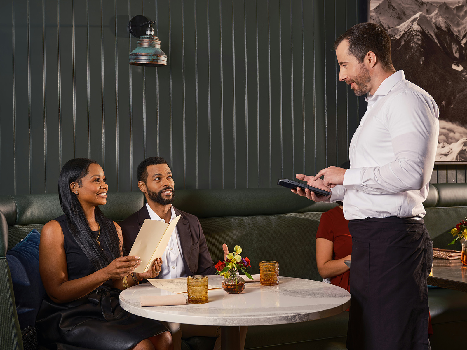 Warm, candid shot of a server taking an order on a handheld at a full table in a busy dining room